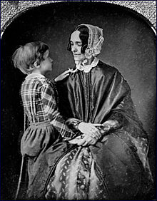 Photographie d'un jeune garçon aux côtés d'une femme assise portant une robe ample