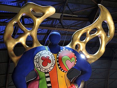 sculpture féminine massive au corps bleu, aux vêtements multicolores et aux ailes dorées, suspendue dans un hall de gare