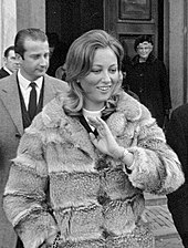 image montrant à l'avant-plan une femme souriante portant un manteau de fourrure devant un homme habillé en civil.