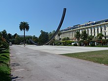 Vue d'une allée de parc dégagée avec palmier et sculpture contemporaine et une façade blanche imposante en fond.