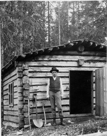 Photo en noir et blanc d'un homme devant une cabane en bois.