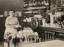 white woman in late 19th-century clothes photographed in a kitchen, with a pile of chickens on the table