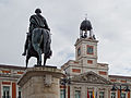 Monument du roi Charles III au centre de la place, devant la Maison royale du courrier et son horloge.