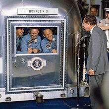 The three crew members smiling at the President through the glass window of their metal quarantine chamber. Below the window is the Presidential Seal, and above it is stenciled on a wooden board "HORNET + 3". President Nixon is standing at a microphone, also smiling. He has dark crinkly hair and a light gray suit.