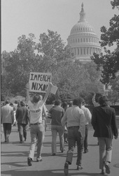 Un manifestant tient au-dessus de sa tête une pancarte où est inscrit « Impeach Nixon ». D'autres personnes marchent comme lui en direction du Capitole.