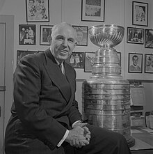 Photo noir et blanc de Clarence Campbell qui pose assis sur le coin d'un bureau à côté de la Coupe Stanley et devant un mur couvert de photos.