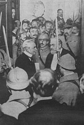 Photo en noir et blanc d’un rassemblement d'individus, avec au centre de l’image deux hommes qui échangent : l’un a une moustache et des cheveux blancs, l’autre présente une chevelure et une barbe grisonnantes et porte des lorgnons