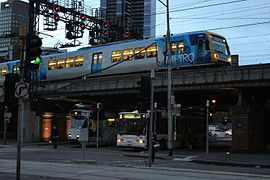 Un tramway à côté d'un bus Melbourne Bus Link, avec un métro Trains X'Trapolis 100 passant au-dessus.