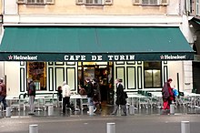 Vue de face d'un café avec son store vert tiré sur lequel est écrit en blanc « Café de Turin » en lettres majuscules.