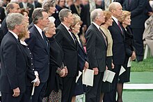 Une rangée de 10 hommes et femmes en couples se tiennent debout lors d'une cérémonie, une foule est présente à l'arrière-plan