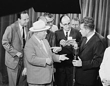 Nixon tient un micro et discute avec un homme portant un chapeau. Plusieurs personnes en costume se tiennent autour des deux hommes.