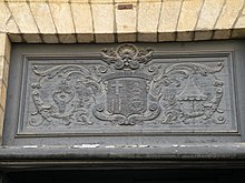 Une photo de l’imposte de la porte principale de l’église. Le blason de la basilique, encadré de ses attributs (pavillon et tintinnabule), est sculpté en bas-relief sur un panneau de bois sombre.