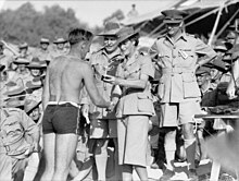 A large crowd of men in uniforms with slouch hats. A woman in uniform with skirt and flat bush hat presents a small trophy to a man in a swim suit.