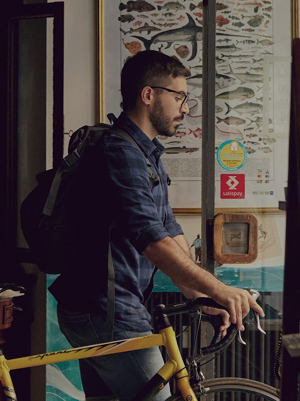 Mattia Garavaglia walks his yellow bike out of his bookstore. He's wearing glasses and a backpack.