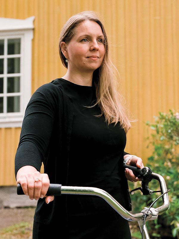 Anne-Christine Hertz, maker van Bikearound, staat voor een geel huis met een fiets. Ze heeft lang haar en draagt ​​een top met lange mouwen.