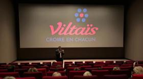 Une salle de cinéma avec des specateurs installés de dos dans des fauteuils rouges, face à un écran affichant le logo de Viltaïs et son slogan Croire en chacun. Un homme seul, face à eux, parle avec un micro dans la main. 