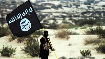 Un homme cagoulé tient un drapeau de l'État islamique dans un désert en Irak ou en Syrie en 2015. (PICTURES FROM HISTORY / UNIVERSAL / GETTY IMAGES)